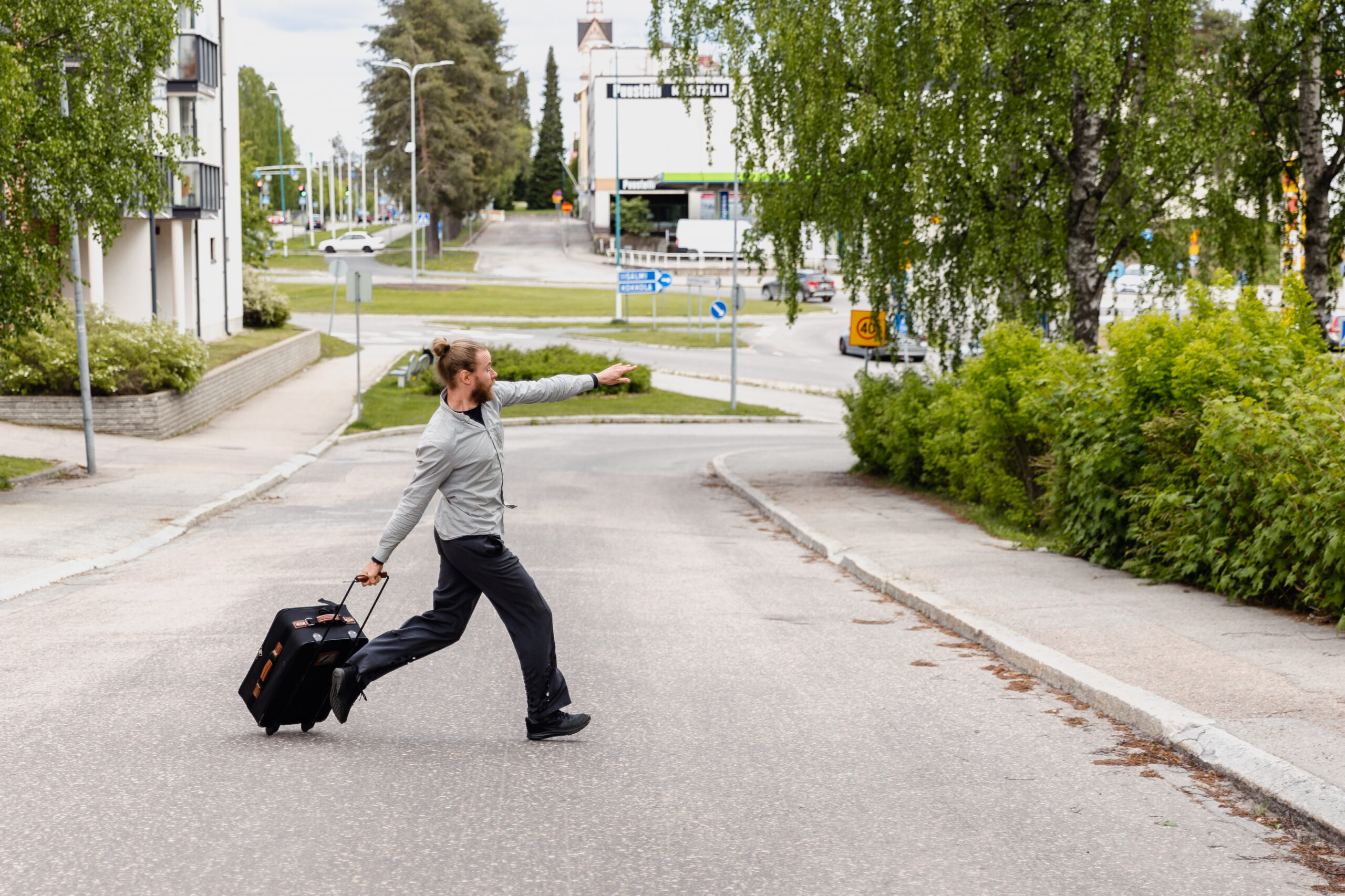 Mies ylittää tien vetäen perässään matkalaukkua. Kuva on Aidatut unelmat Kajaani -esityksestä.